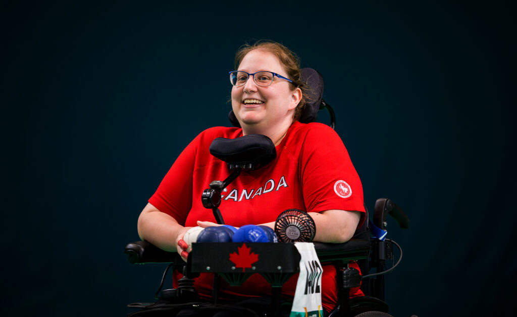La joueuse canadienne de boccia Alison Levine sourit pendant une compétition aux Jeux parapanaméricains de Santiago 2023. Elle porte un chandail rouge de l’équipe canadienne et est assise dans son fauteuil roulant motorisé avec des balles de boccia et un plateau d’équipement devant elle.