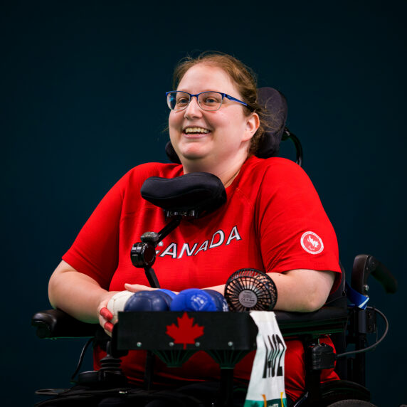 La joueuse canadienne de boccia Alison Levine sourit pendant une compétition aux Jeux parapanaméricains de Santiago 2023. Elle porte un chandail rouge de l’équipe canadienne et est assise dans son fauteuil roulant motorisé avec des balles de boccia et un plateau d’équipement devant elle.