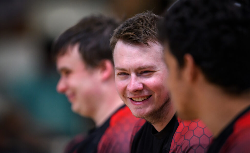 L’athlète canadien de goalball Blair Nesbitt sourit assis sur le banc avec ses coéquipiers pendant un match aux Jeux parapanaméricains de Santiago 2023. Il porte le maillot rouge et noir de l’équipe du Canada.