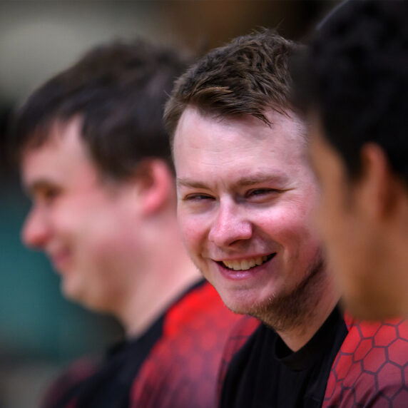 L’athlète canadien de goalball Blair Nesbitt sourit assis sur le banc avec ses coéquipiers pendant un match aux Jeux parapanaméricains de Santiago 2023. Il porte le maillot rouge et noir de l’équipe du Canada.