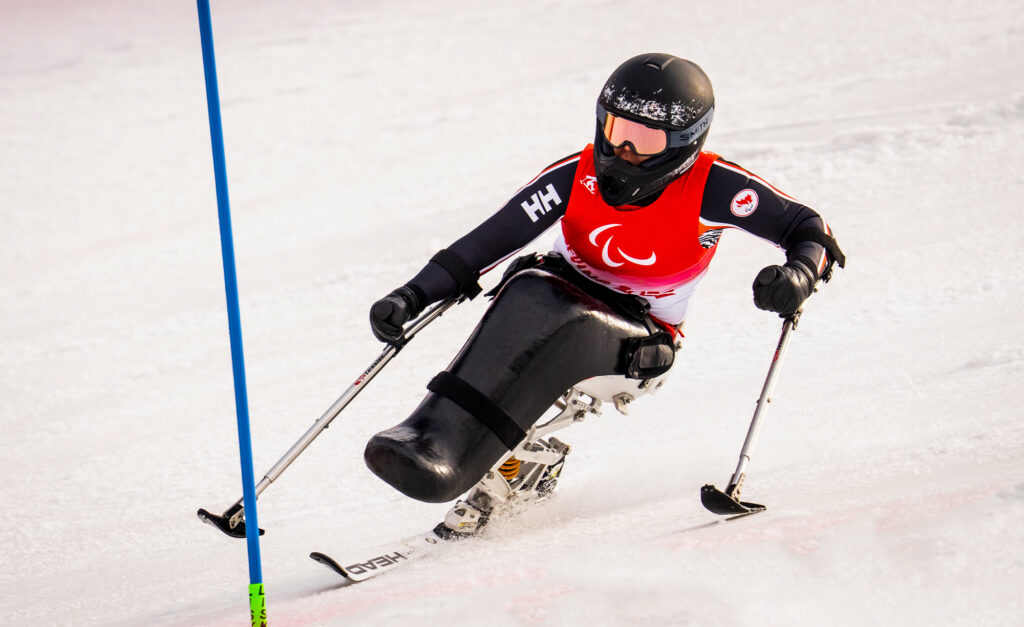 La paralympienne canadienne Katie Combaluzier participe à l’épreuve de slalom aux Jeux paralympiques d’hiver de Beijing 2022, franchissant une porte au Centre national de ski alpin de Yanqing, dans un monoski noir, vêtue d’un dossard rouge et d’un casque noir.