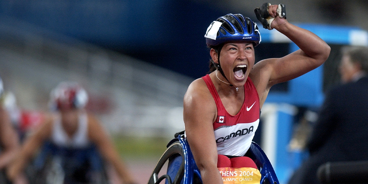 La Paralympienne canadienne Chantal Petitclerc célèbre avec un puissant geste du poing après avoir remporté une course en fauteuil roulant aux Jeux paralympiques d’Athènes 2004. Portant l’uniforme rouge et blanc d’Équipe Canada et un casque, elle crie de joie et de détermination en franchissant la ligne d’arrivée.