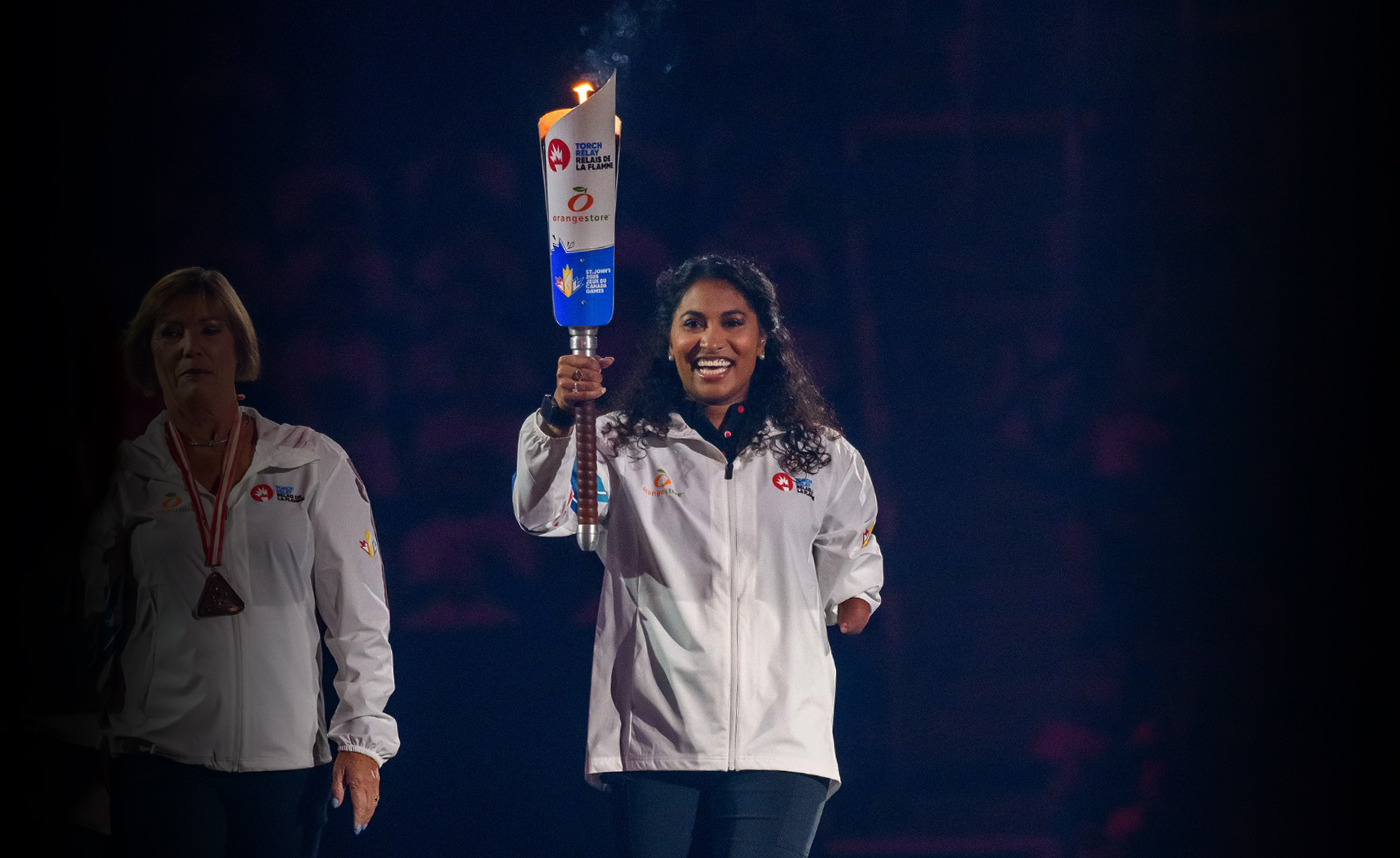 La paranageuse canadienne Katarina Roxon sourit en tenant fièrement la flamme allumée lors de la cérémonie d’ouverture des Jeux du Canada 2025. Elle porte une veste blanche de l’équipe de Terre-Neuve-et-Labrador.