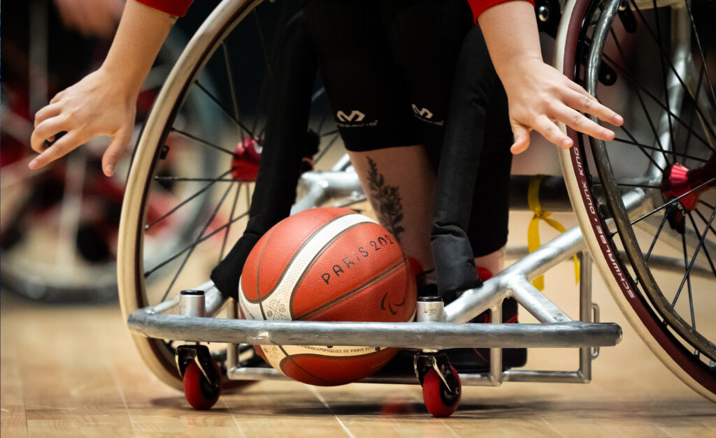 Gros plan d’un match de basketball en fauteuil roulant. Une main de joueur tendue vers le ballon rouge et blanc marqué « Paris 2024 » sur un plancher de bois. Dynamique et concentré.       Ask ChatGPT