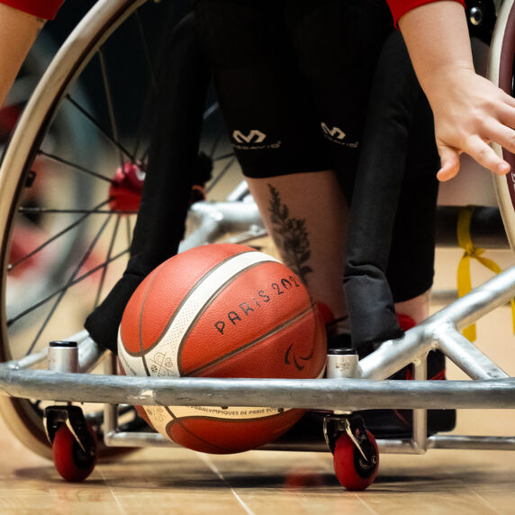 Gros plan d’un match de basketball en fauteuil roulant. Une main de joueur tendue vers le ballon rouge et blanc marqué « Paris 2024 » sur un plancher de bois. Dynamique et concentré.       Ask ChatGPT