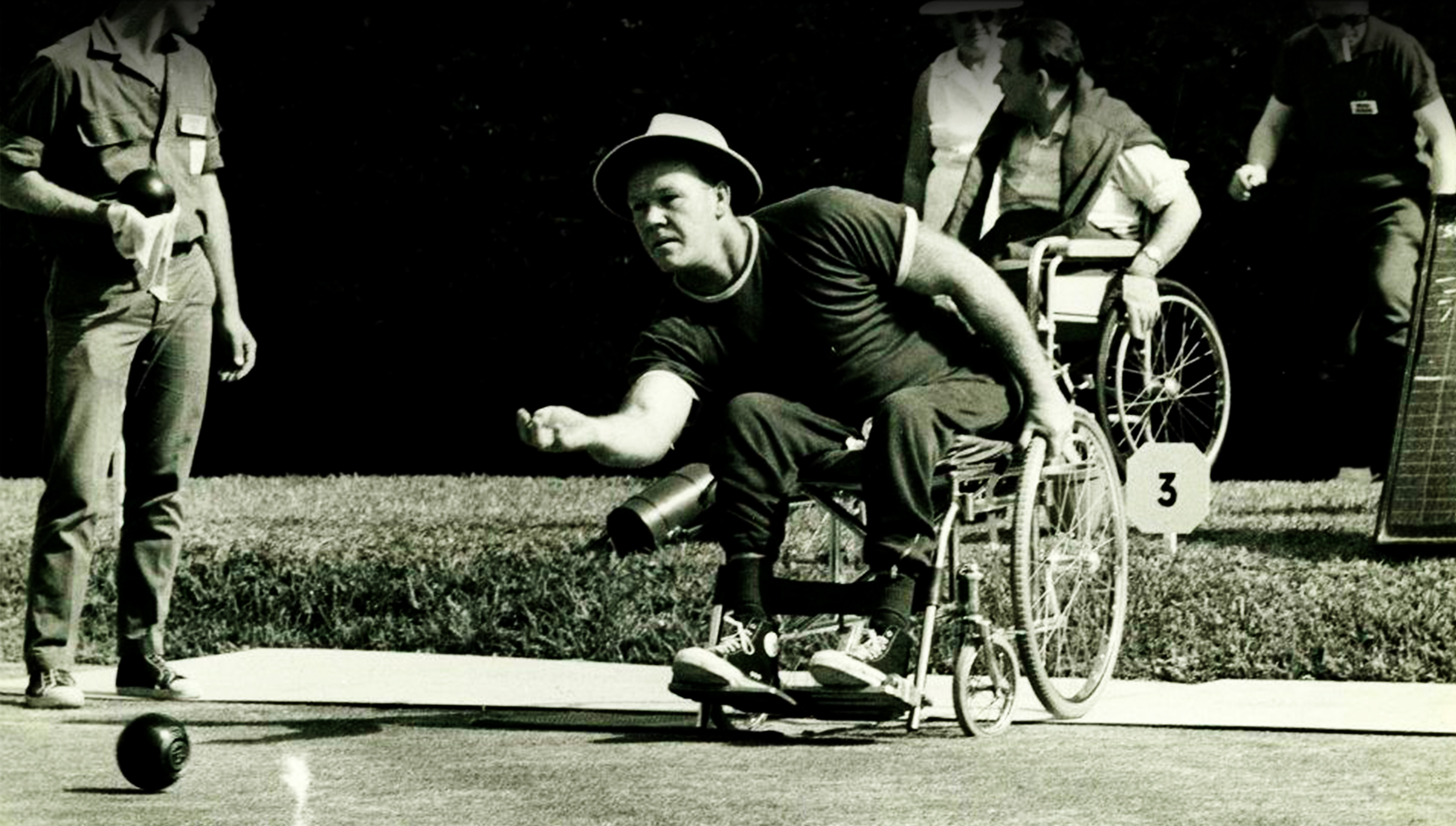 Photo en noir et blanc d’un athlète de boulingrin paralympique aux Jeux paralympiques de Tel-Aviv 1968. L’athlète, assis dans un fauteuil roulant et portant un chapeau et un chandail foncé, est en plein lancer sur un terrain de boulingrin. D’autres participants et officiels observent à l’arrière-plan.