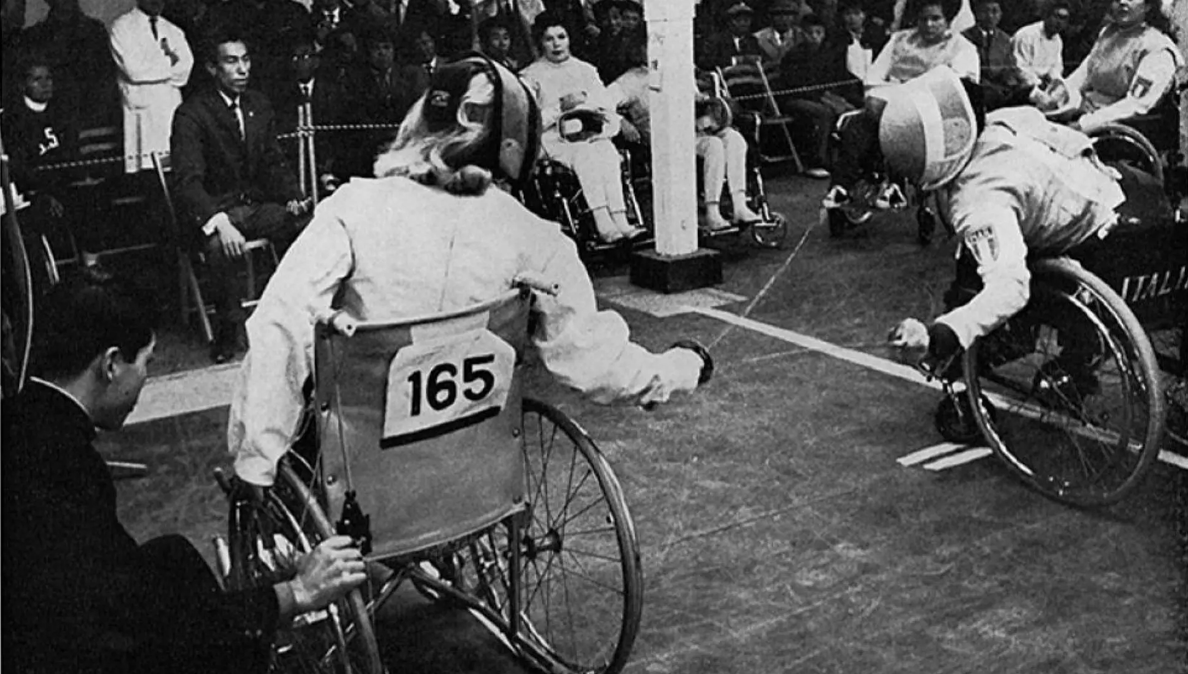 Photo en noir et blanc d’un match d’escrime en fauteuil roulant aux Jeux paralympiques de Tokyo 1964. Deux athlètes en fauteuil roulant, portant un équipement de protection, s’affrontent. L’athlète numéro 165 est à gauche, et son adversaire italien à droite. Un public observe en arrière-plan.