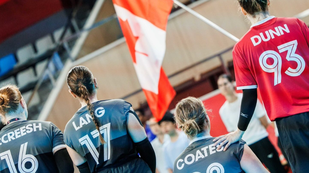 Des joueuses canadiennes de volleyball assis, Ecreesh (16), Lang (17) et Coates, se tiennent ensemble face au drapeau canadien, aux côtés d'un joueur d'Équipe Canada portant le chandail « Dunn 93 » dans l'aréna.